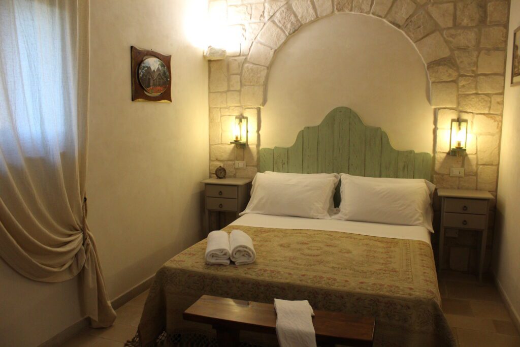 descrizione-queen-1 Camera da letto in stile rustico con letto matrimoniale, testiera in legno chiaro, pareti in pietra, due comodini con lampade, un quadro incorniciato e una tenda chiara.