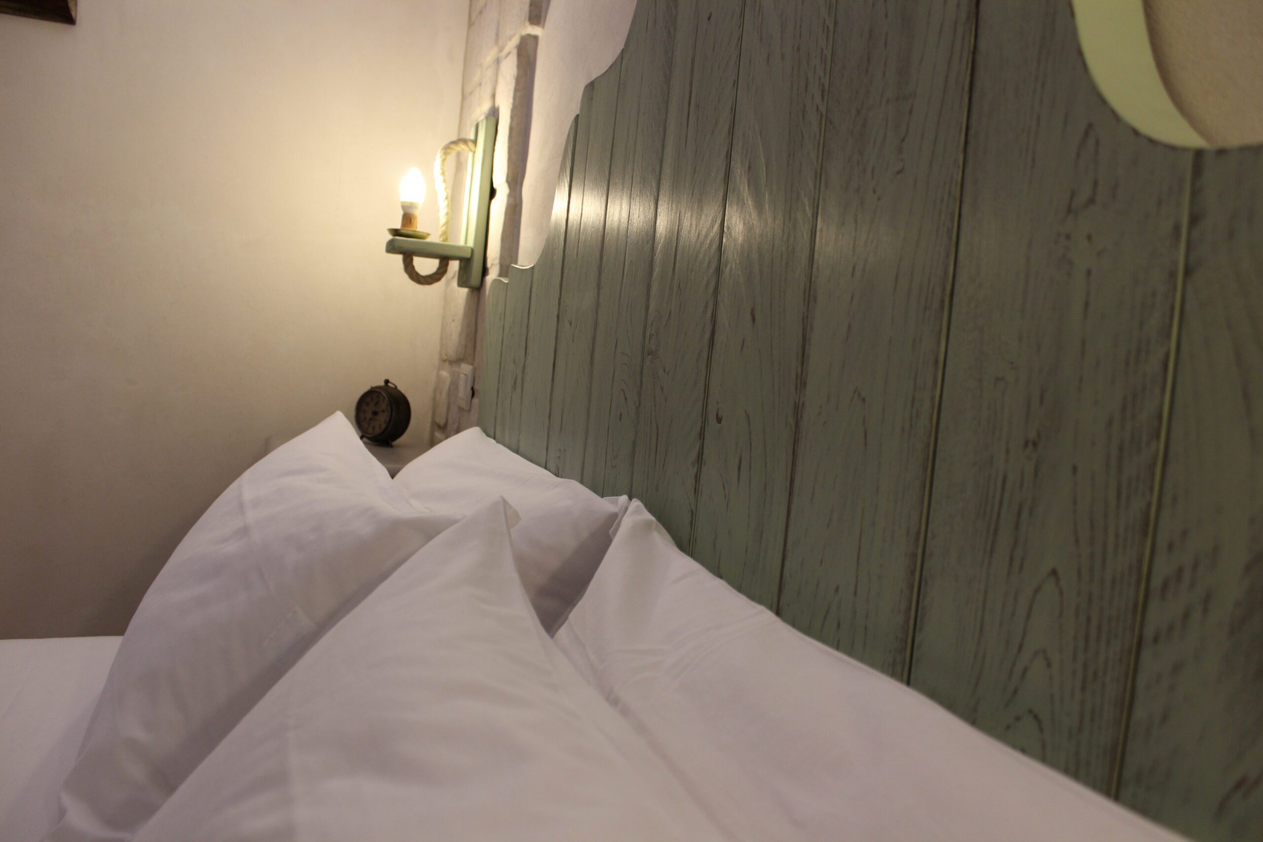 descrizione-queen-2 Un letto con cuscini bianchi e testiera in legno chiaro, con una lampada da parete accesa e una sveglia su un comodino.