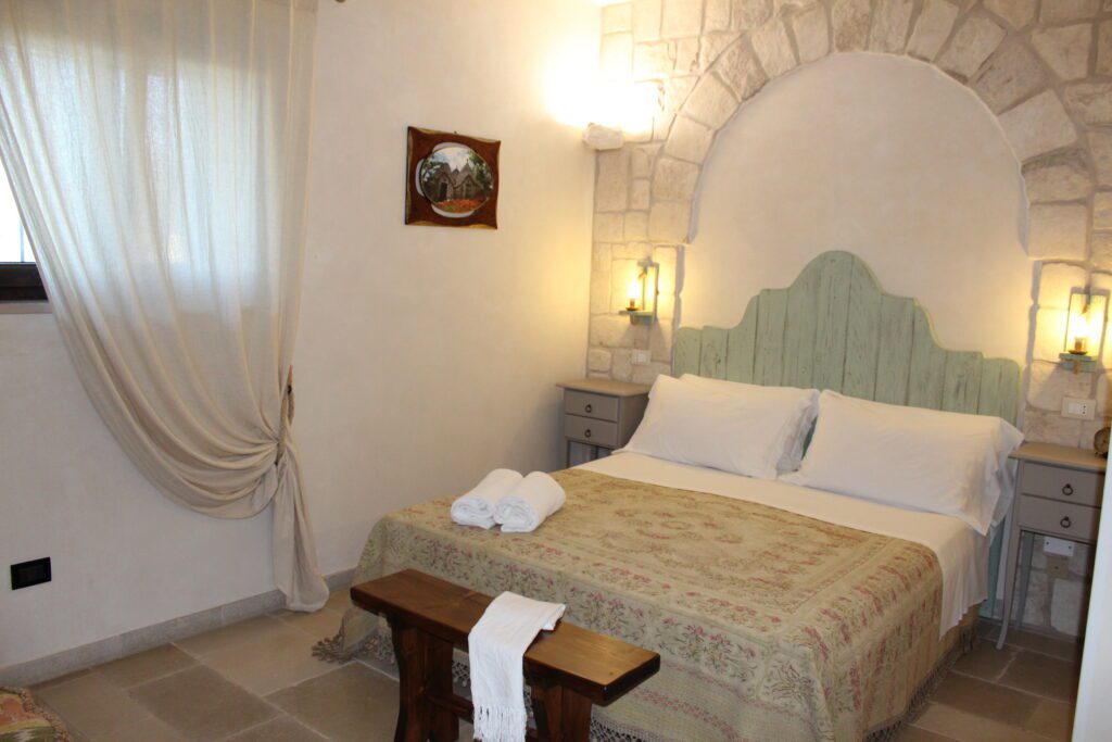 descrizione-queen-3 Camera da letto con letto matrimoniale, testiera in legno chiaro, pareti in pietra, due comodini con lampade, un quadro incorniciato, una finestra con tende e un baule in legno ai piedi del letto.
