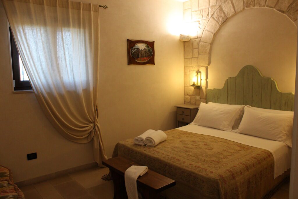 descrizione-queen-4 Camera da letto con letto matrimoniale, testiera in legno chiaro, pareti in pietra, due comodini con lampade, un quadro incorniciato, una finestra con tende e un baule in legno ai piedi del letto.