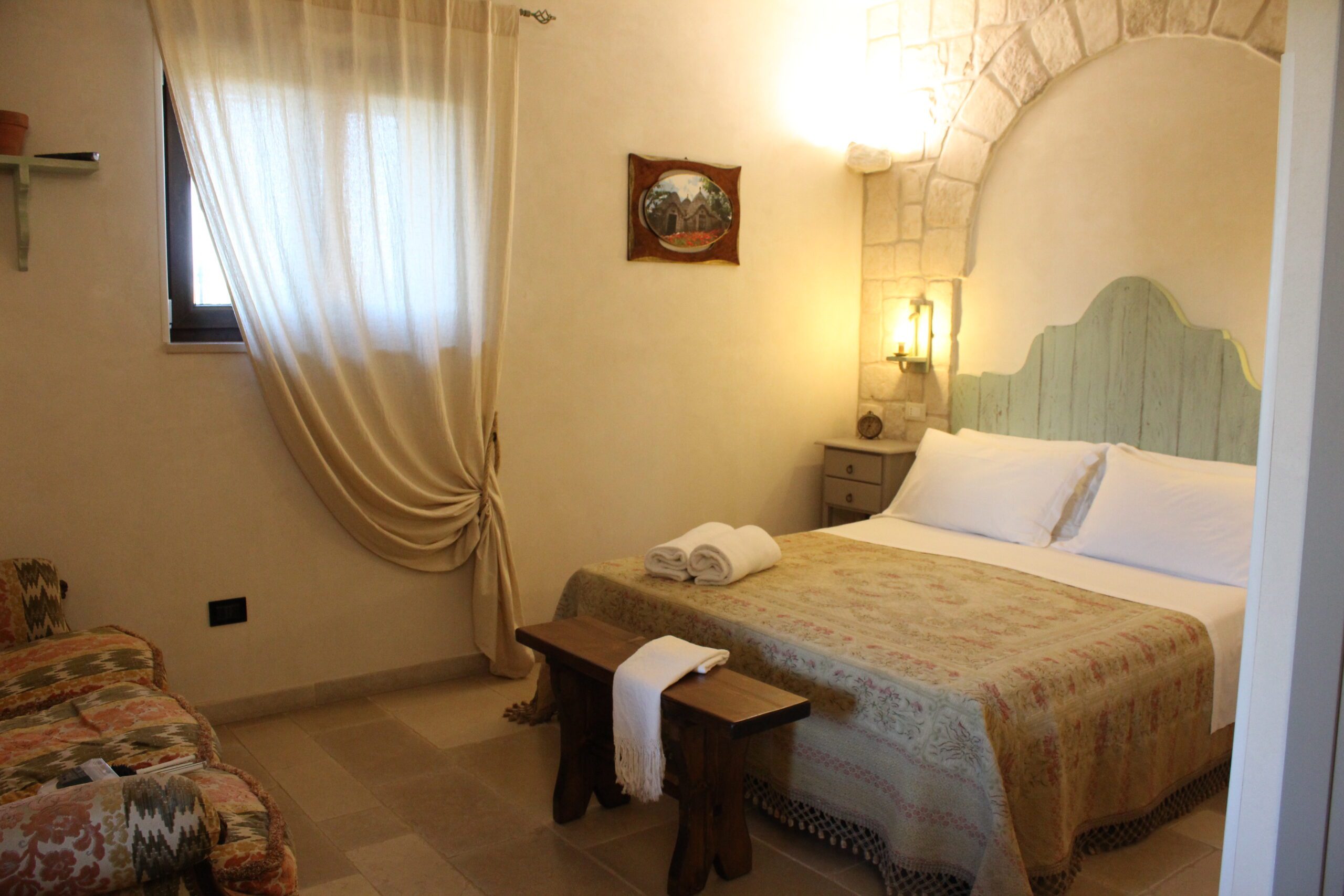 Camera da letto con letto matrimoniale, testiera in legno chiaro, pareti in pietra, due comodini con lampade, un quadro incorniciato, una finestra con tende e un divano sulla sinistra.