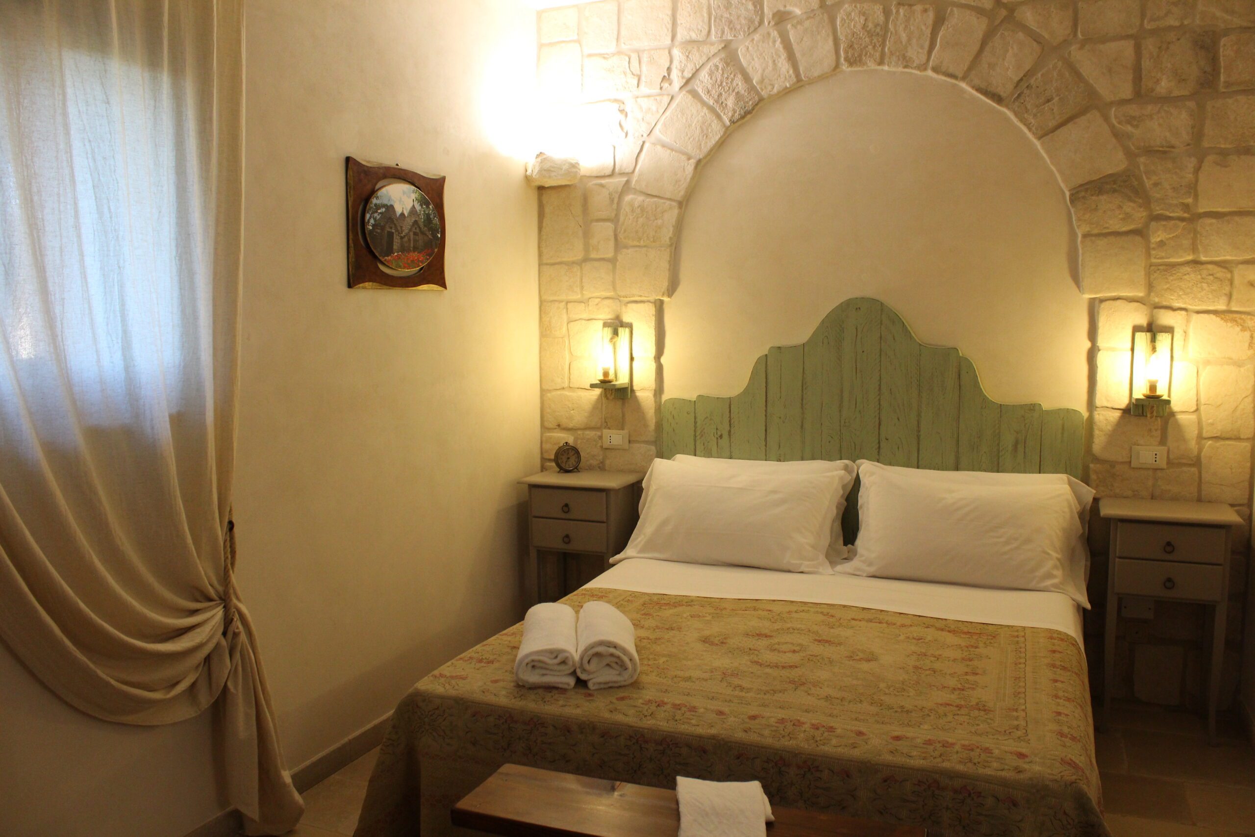 Camera da letto con letto matrimoniale, testiera in legno chiaro, pareti in pietra, due comodini con lampade, un quadro incorniciato, una finestra con tende e un divano sulla sinistra.