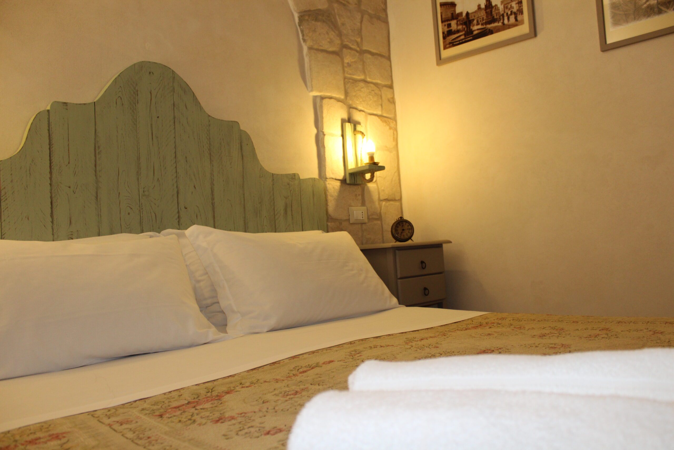 Letto matrimoniale con testiera in legno chiaro, cuscini bianchi, copriletto fantasia, comodini con lampade accese e parete in pietra rustica in una camera da letto accogliente.